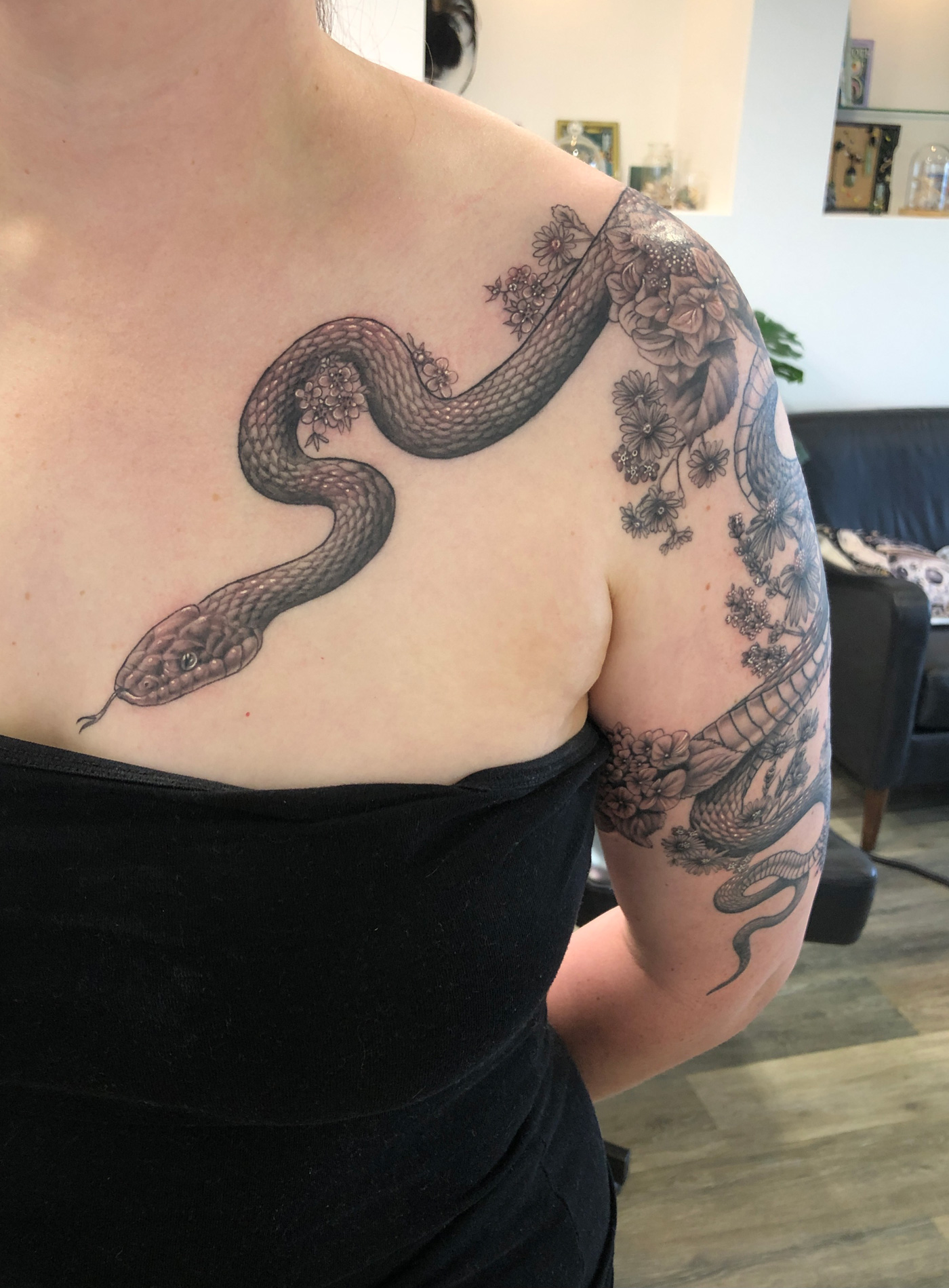Black and grey snake tattoo entwined with wildflowers and hydrangeas wrapping around a woman's arm and flowing onto the chest.