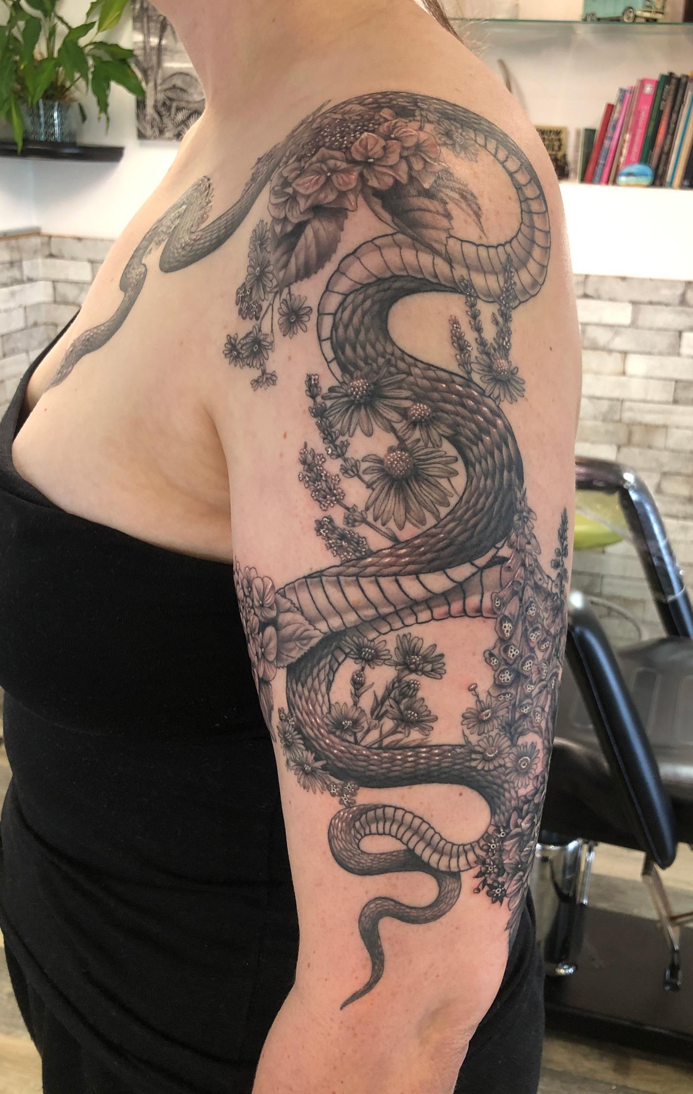 Black and grey snake tattoo weaving through delicate wildflowers and hydrangeas across a woman's shoulder, arm and chest.
